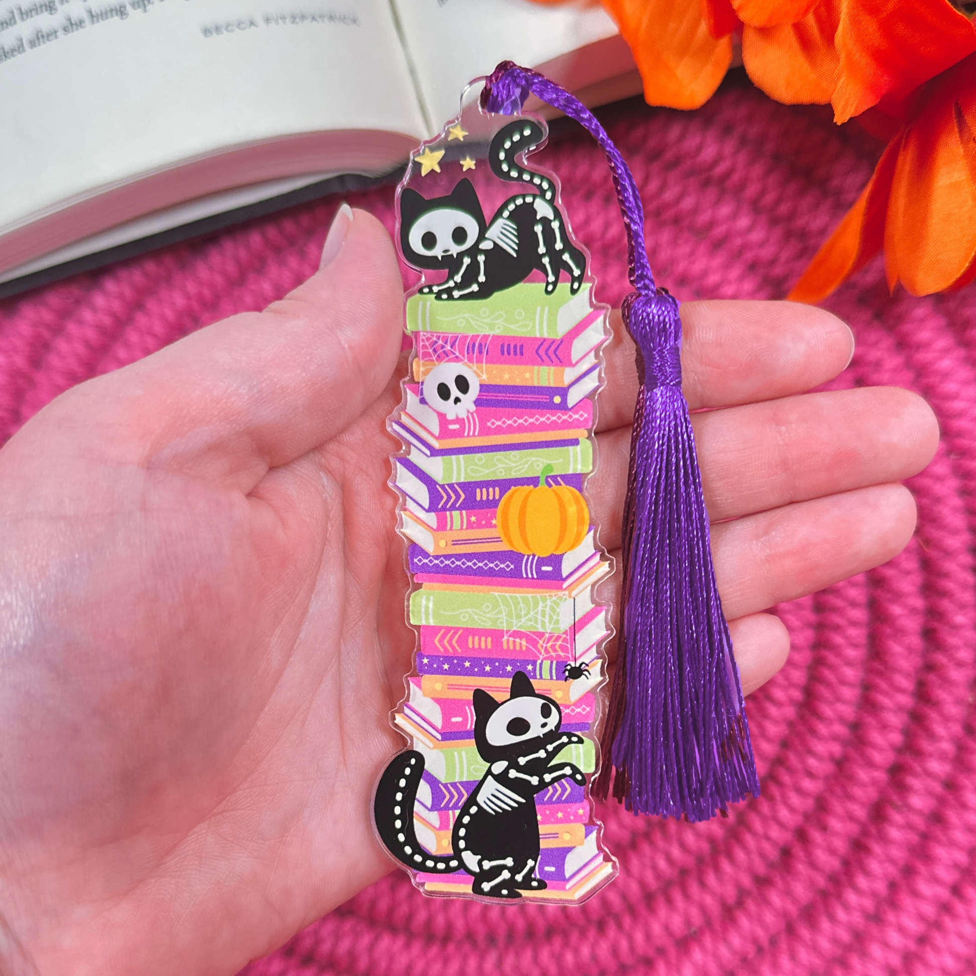 Colorful bookmark with a Halloween cat design held by a hand, with flowers and an open book in the background.