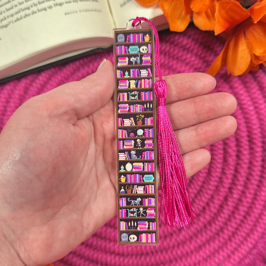 Hand holding a decorative bookmark with tassel on a pink woven surface with orange flowers.