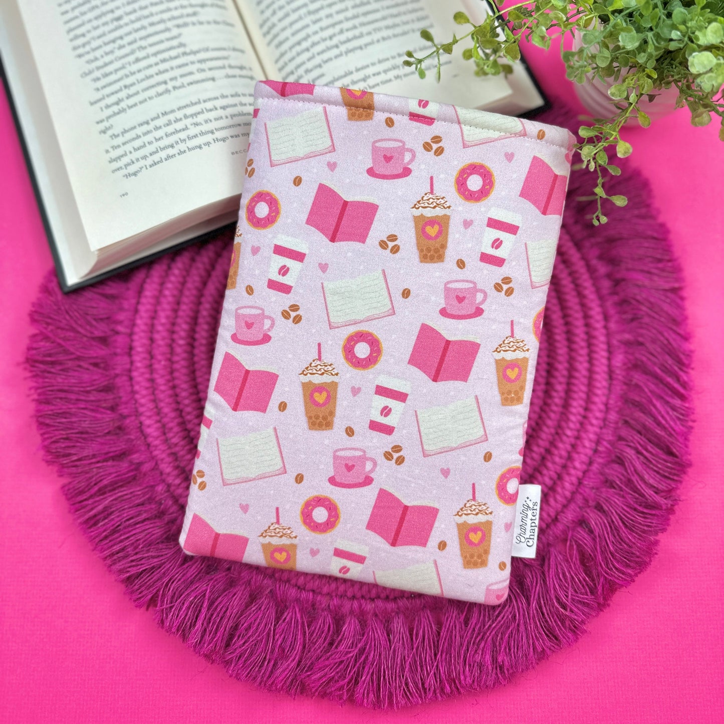 Pink fabric book cover with a pink coffee and donut themed design on a pink surface with an open book and plant.