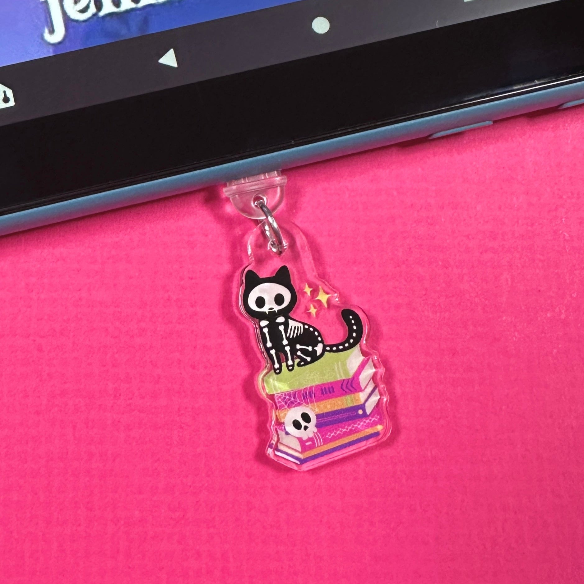 Halloween-themed dust plug charm with a skeleton cat sitting on a bookstack.  The charm is displayed in a blue Kindle which is laying n a hot pink surface.