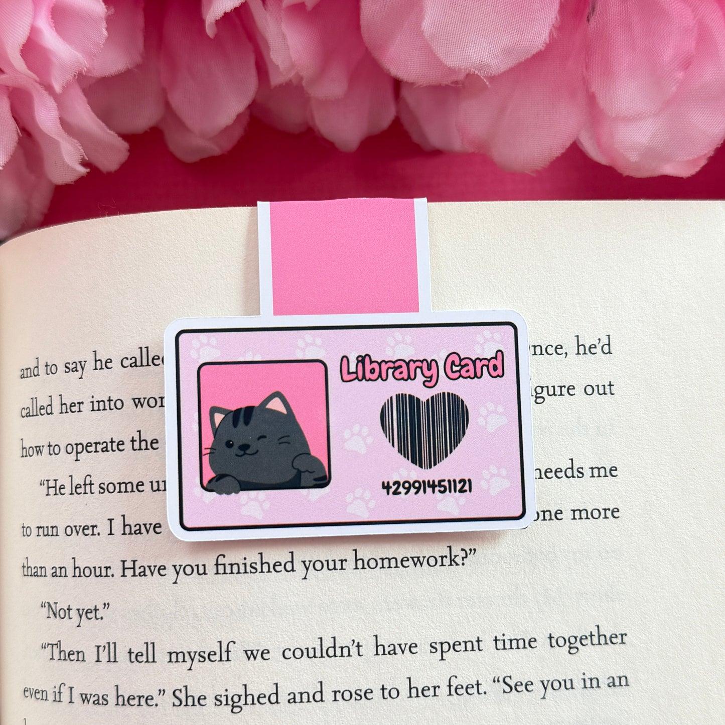 Magnetic bookmark that has a design of a pink library card with a black cat. The bookmark is on an open book surrounded by pink flowers.