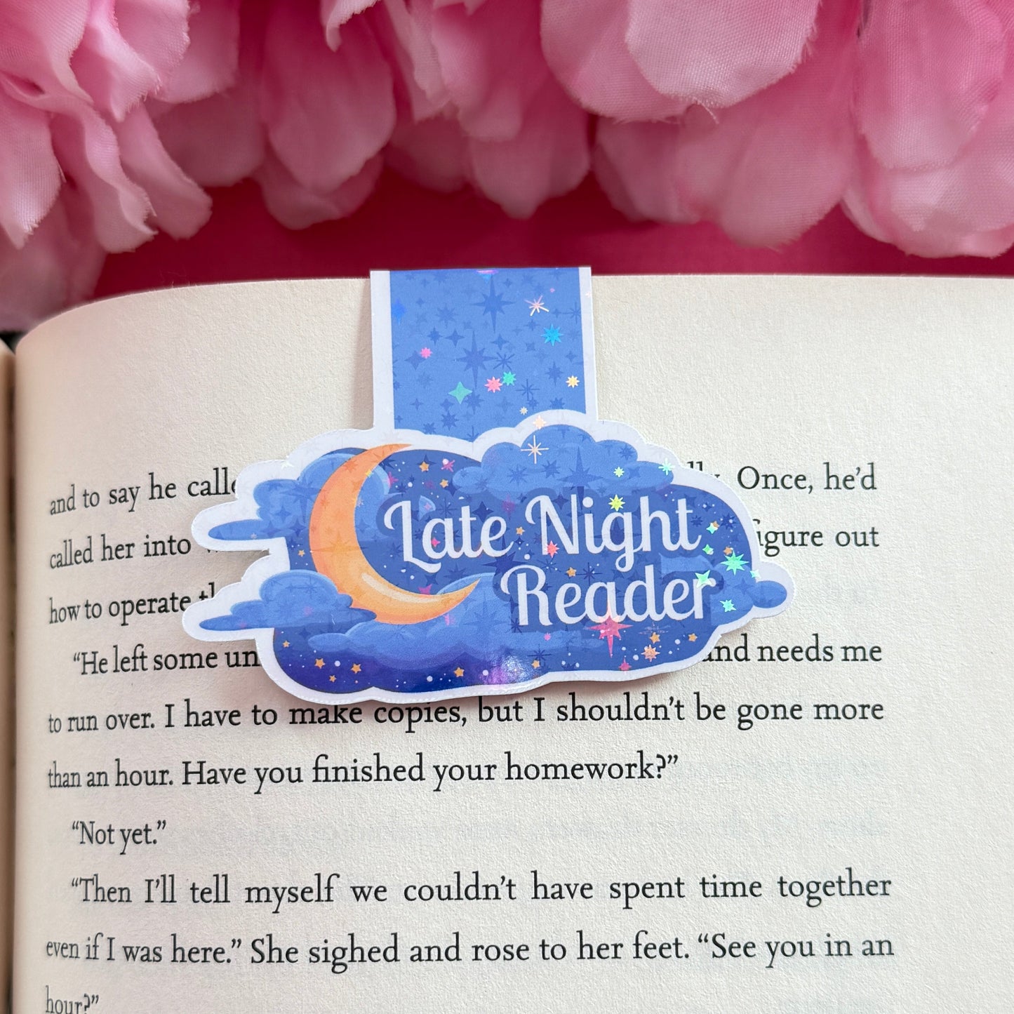 Magnetic bookmark that has a later night read design. The bookmark is on an open book surrounded by pink flowers.