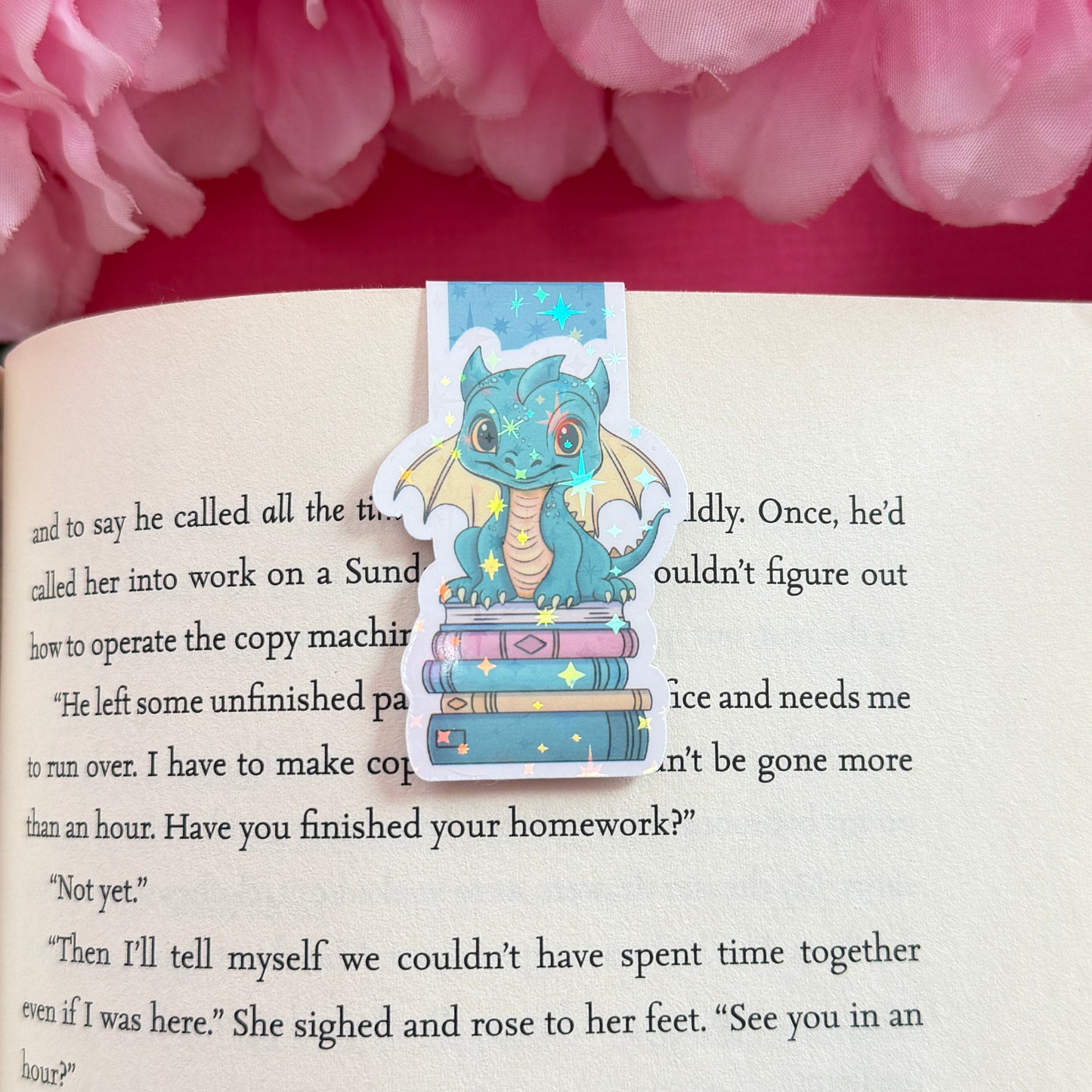 Magnetic bookmark with a teal and yellow dragon design on an open book surrounded by pink flowers. The bookmark has a holographic star finish.