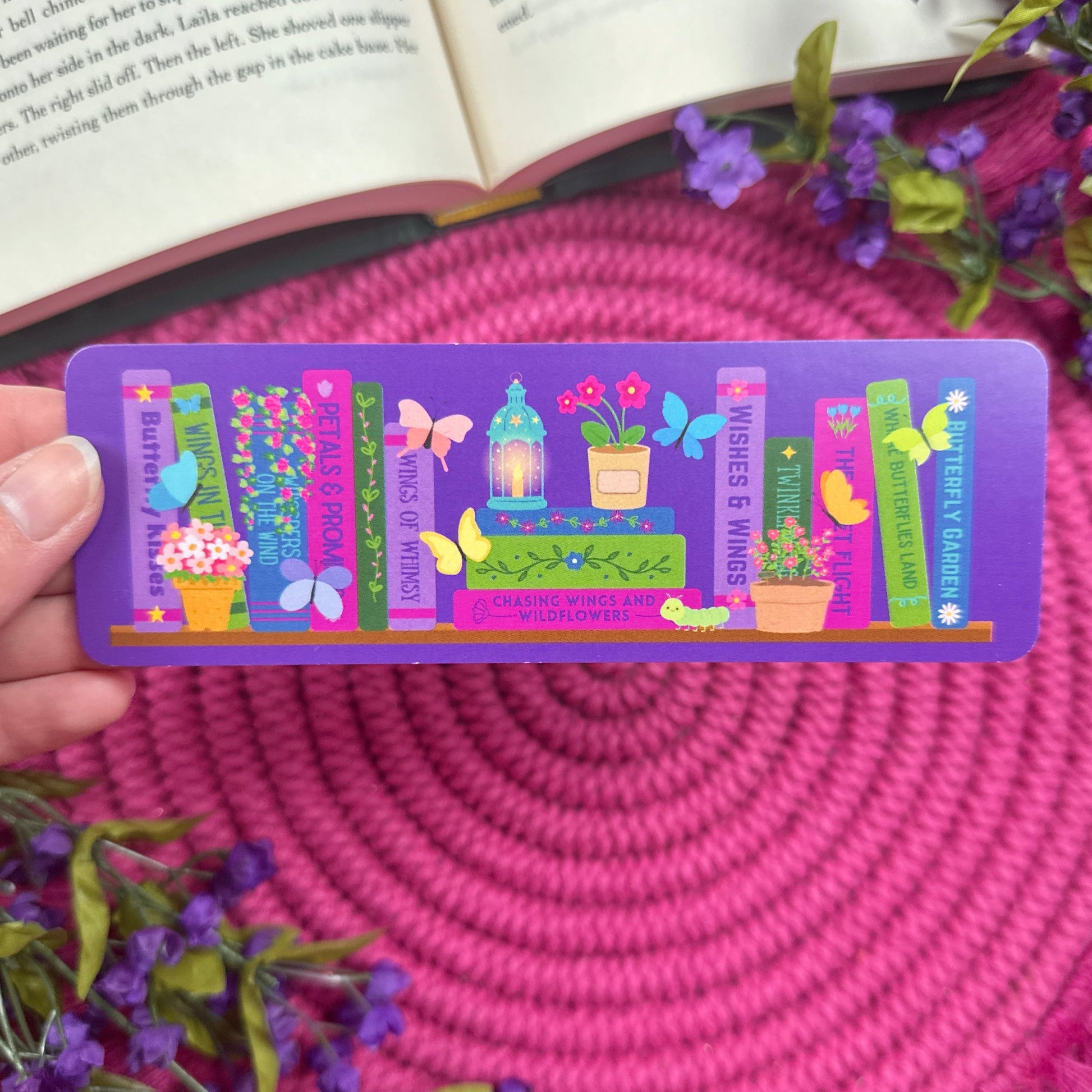 Colorful bookmark with butterflies on a bookshelf with an enchanted garden theme. Hand is holding bookmark in front of a book laying on a pink woven mat with purple flowers around book.