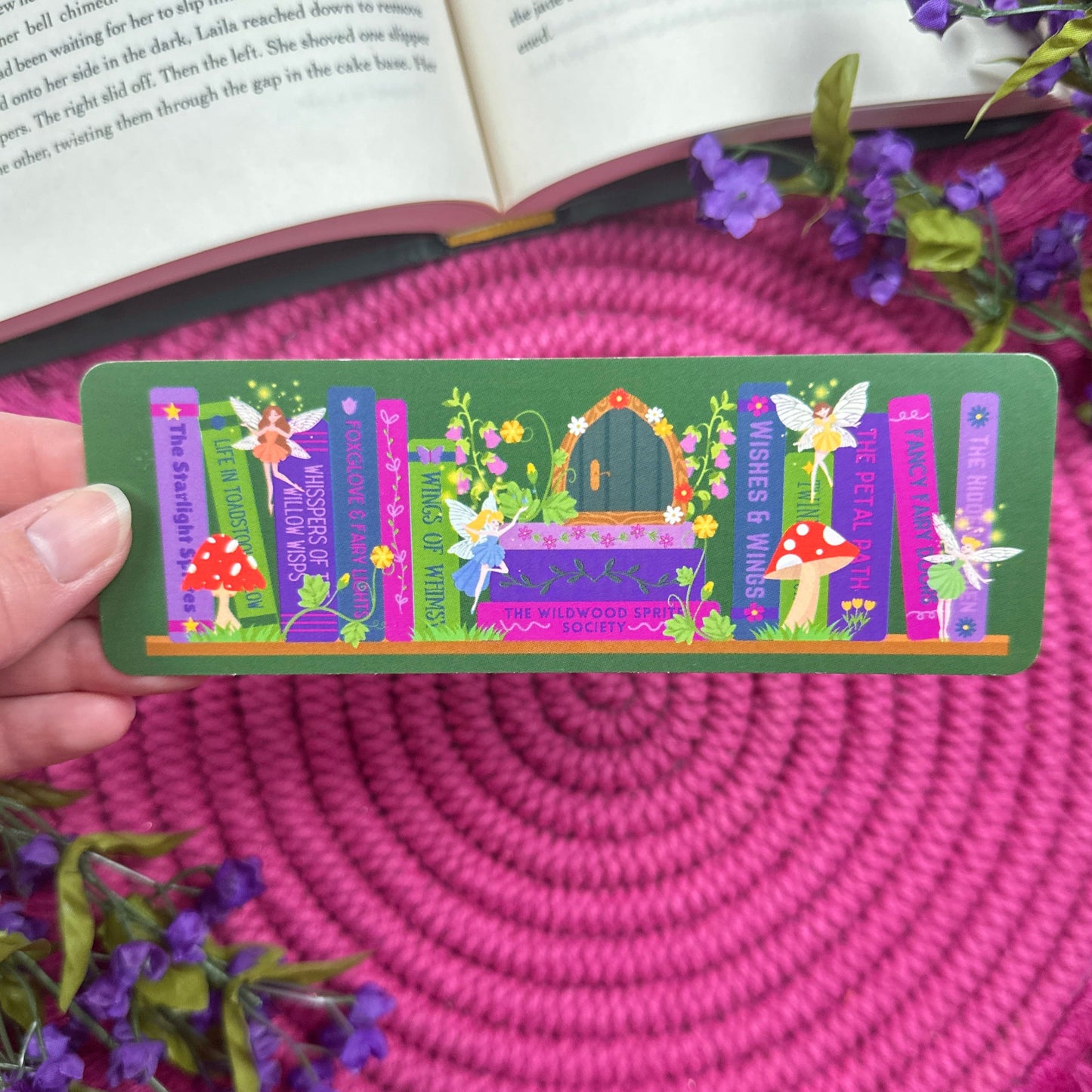 Colorful bookmark with a frog sitting on a bookshelf with an enchanted garden theme. Hand is holding bookmark in front of a book laying on a pink woven mat with purple flowers around book.