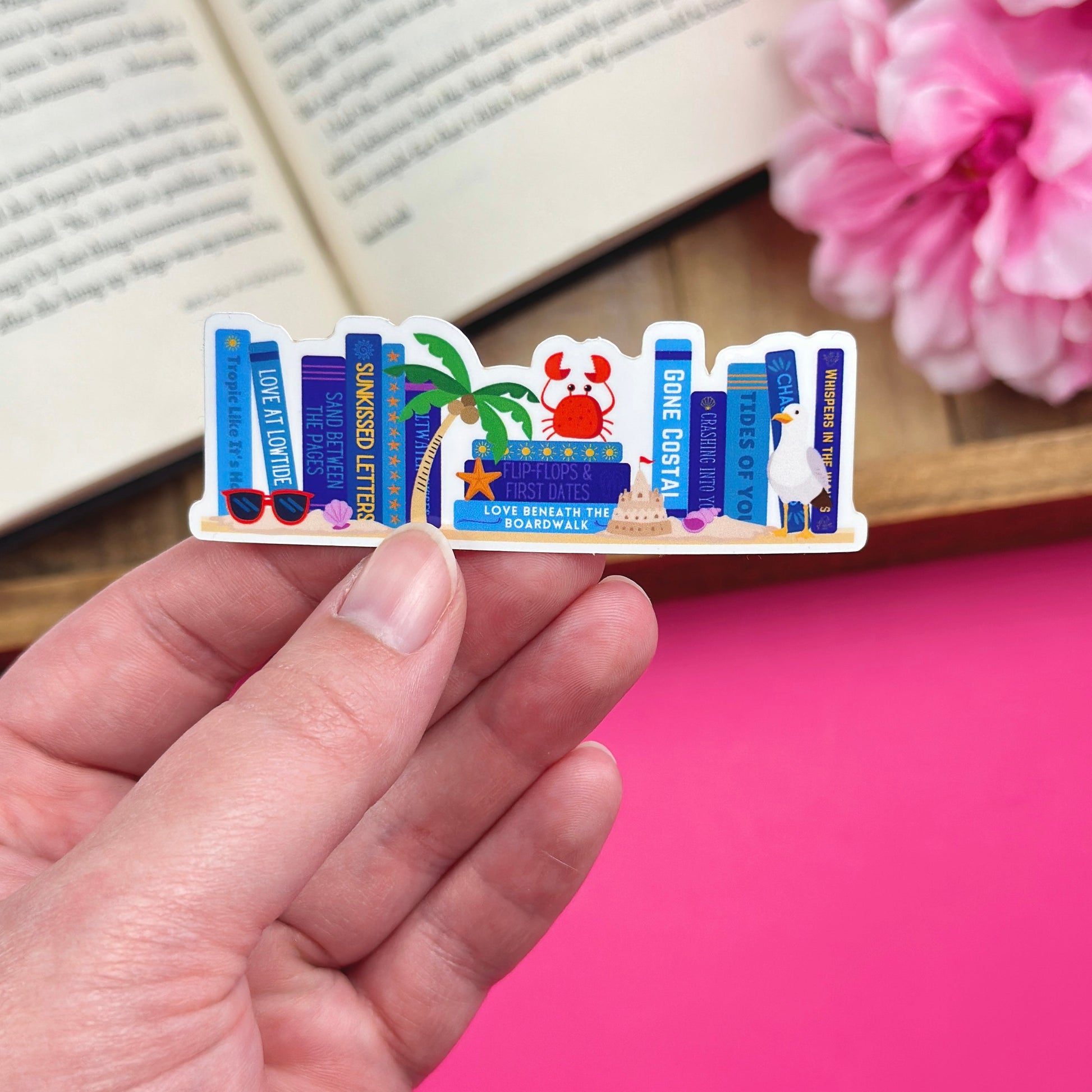 Hand holding a vinyl sticker with a beach bookshelf design, with an open book and pink flowers in the background.