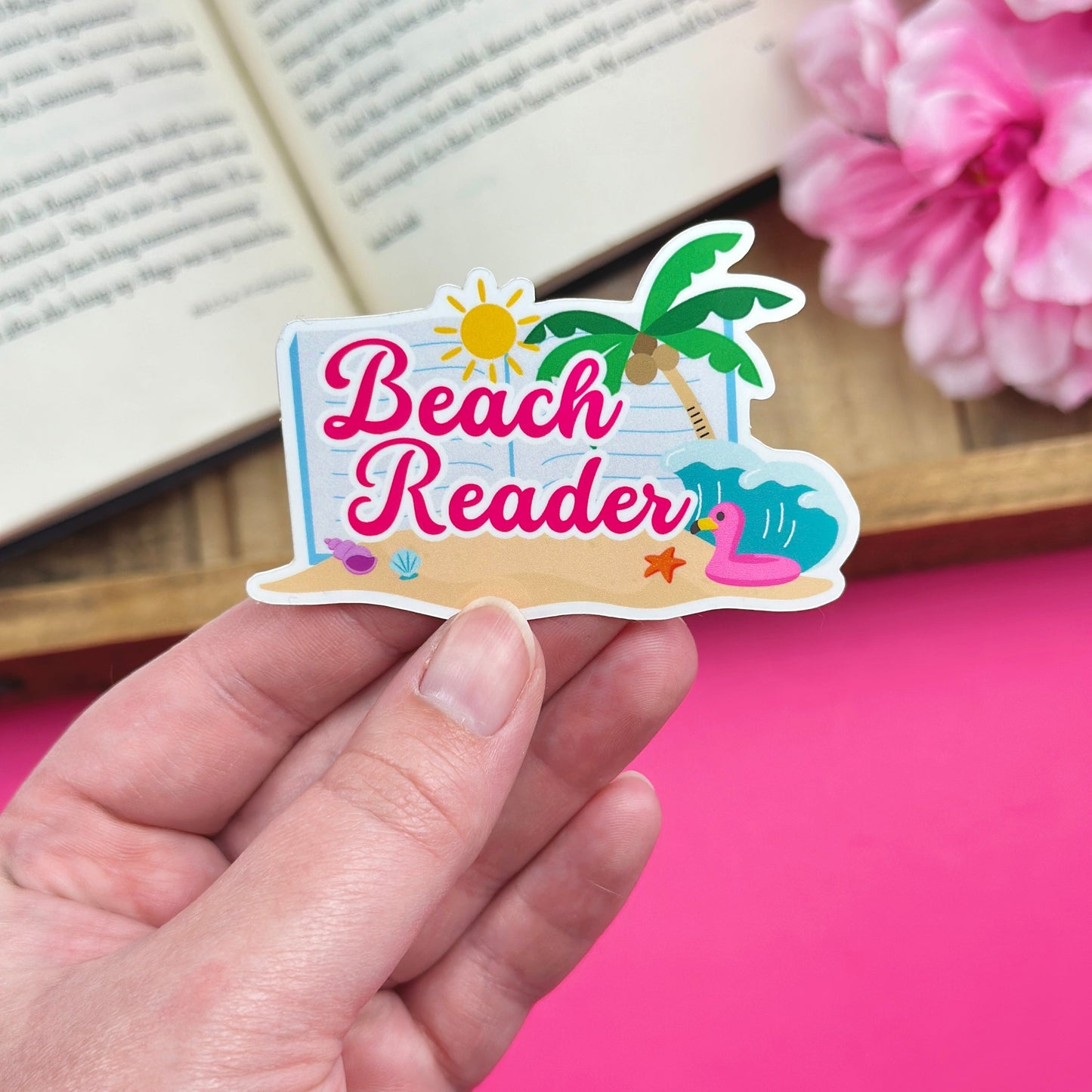 Hand holding a sticker with a beach reader design, with an open book and flowers in the background.