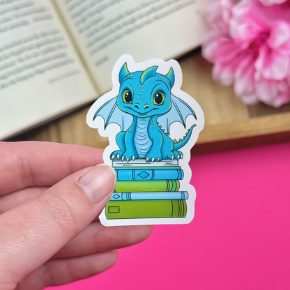 Hand holding a vinyl sticker with a blue and green dragon sitting atop a bookstack, with an open book and pink flowers in the background.
