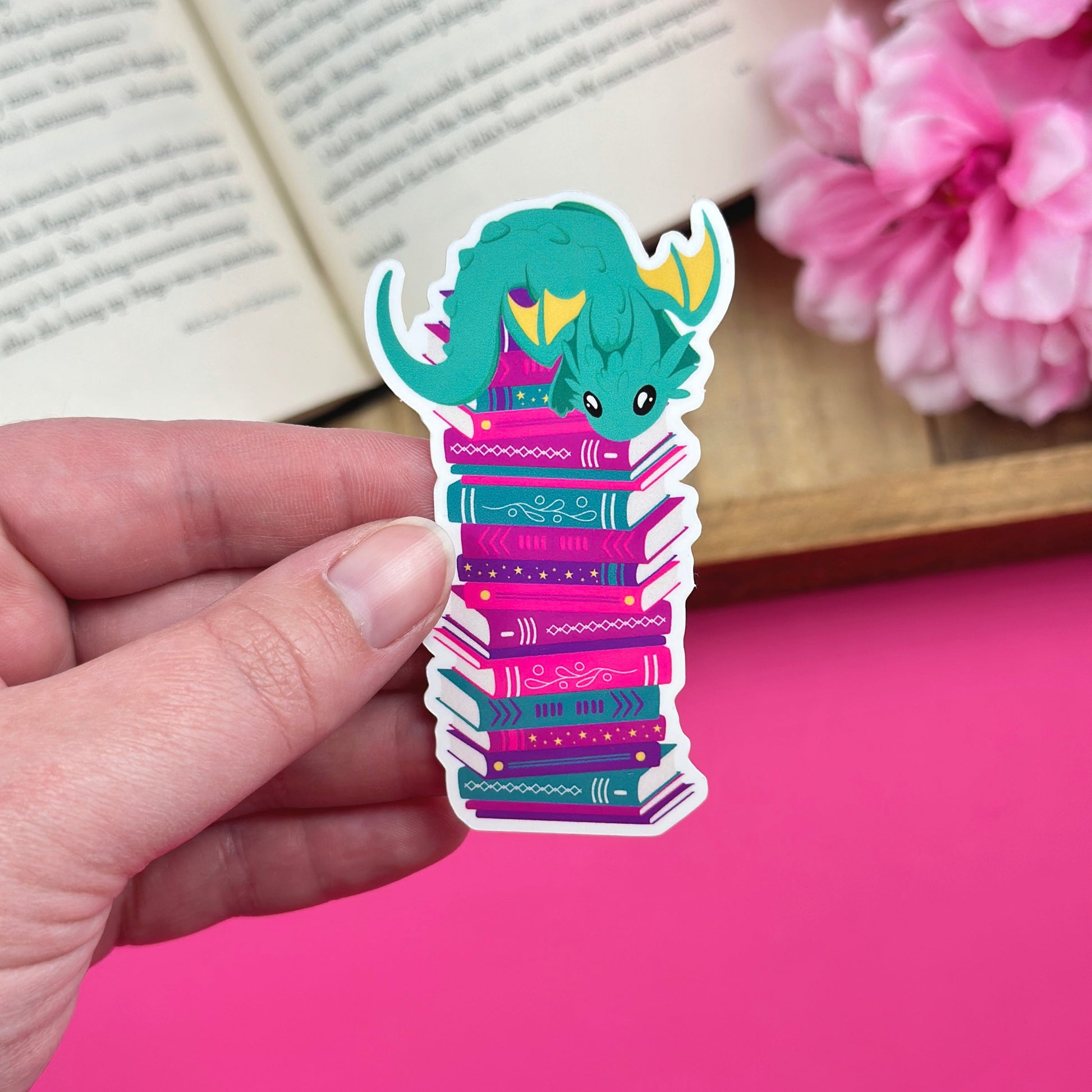 Hand holding a vinyl sticker of a green dragon sitting on a stack of books, with an open book and pink flowers in the background.