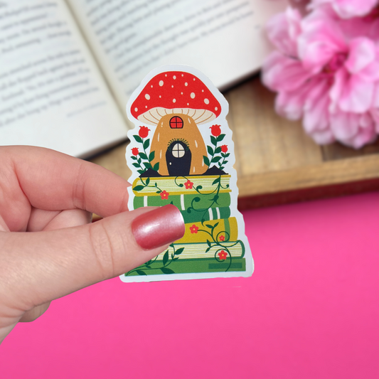 Hand holding a vinyl sticker with a cottagecore mushroom house sitting atop a bookstack, with an open book and pink flowers in the background.