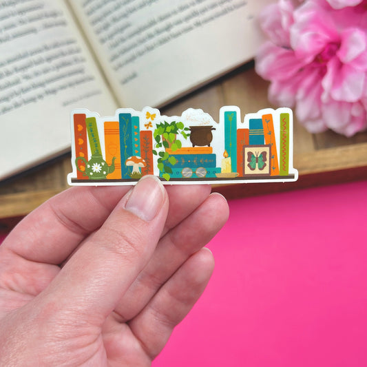 Hand holding a sticker with a cottagecore bookshelf design, with an open book and flowers in the background