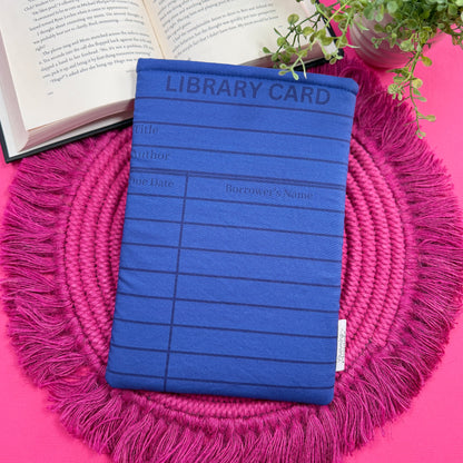 Padded book sleeve with a blue library card design on a pink surface with an open book and plant in the background.