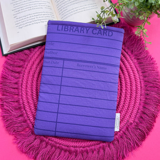Padded book sleeve with a purple library card design on a pink surface with an open book and plant in the background.