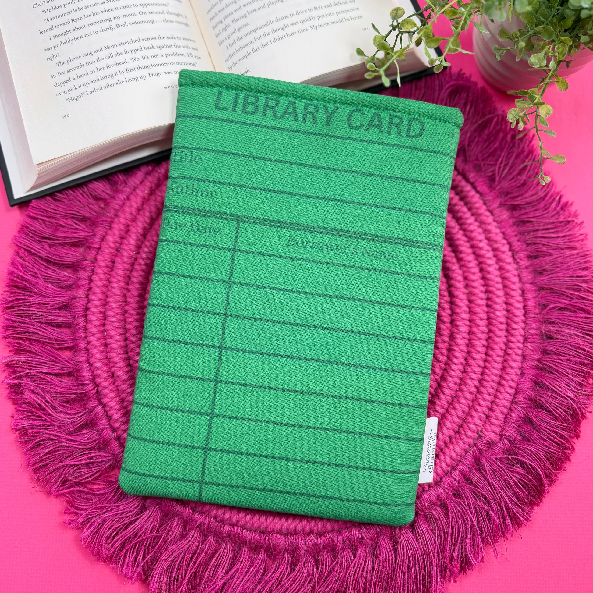Padded book sleeve with a green library card design on a pink surface with an open book and plant in the background.