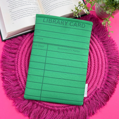 Padded book sleeve with a green library card design on a pink surface with an open book and plant in the background.
