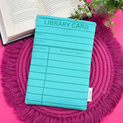 Padded book sleeve with a turquoise blue library card design on a pink surface with an open book and plant in the background.