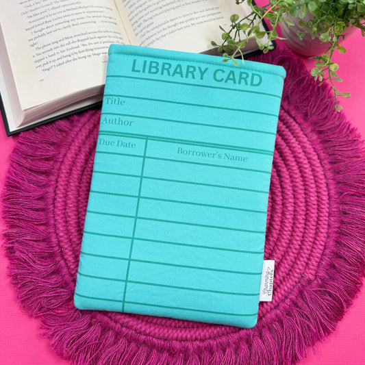 Padded book sleeve with a turquoise blue library card design on a pink surface with an open book and plant in the background.