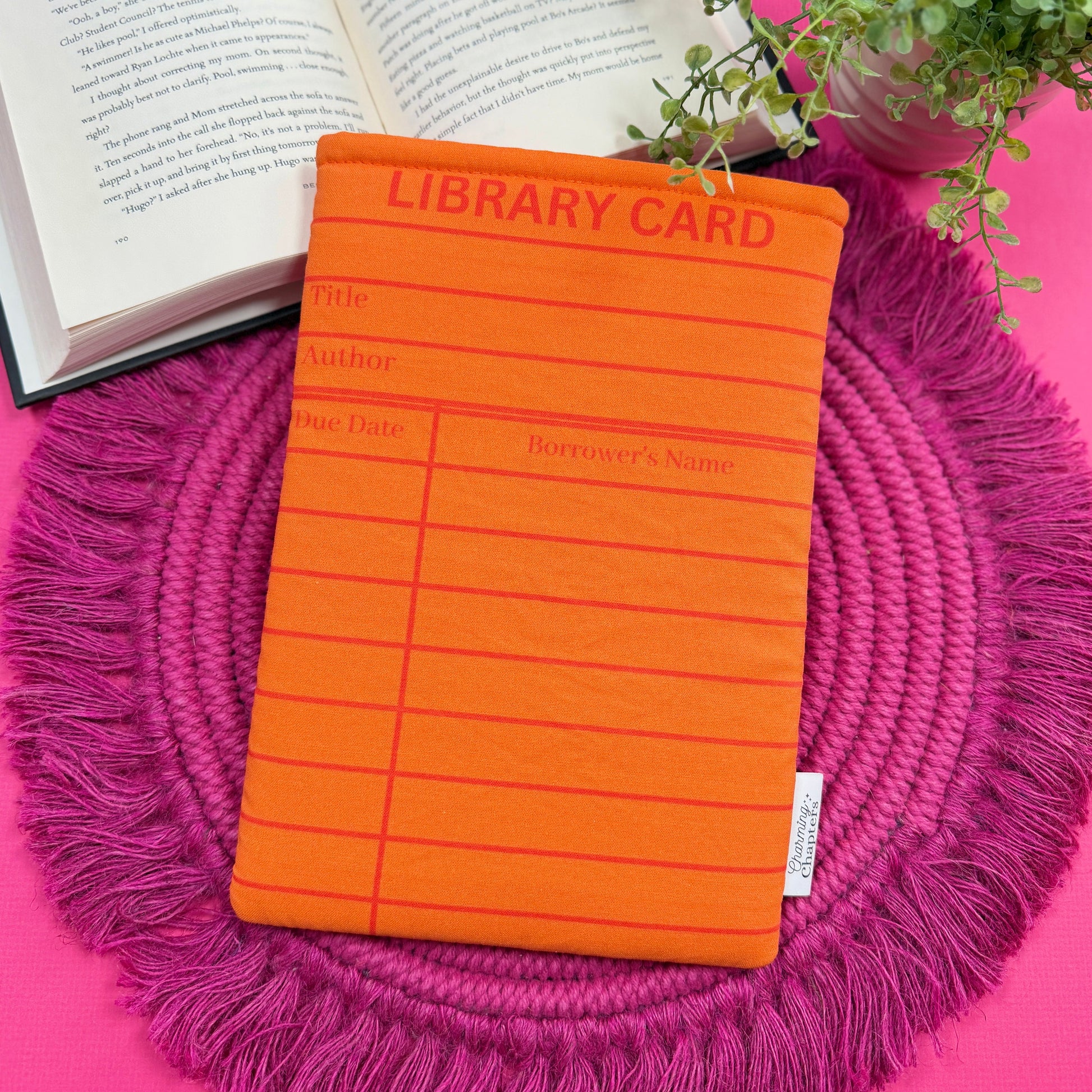 Padded book sleeve with an orange library card design on a pink surface with an open book and plant in the background.