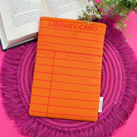 Padded book sleeve with an orange library card design on a pink surface with an open book and plant in the background.