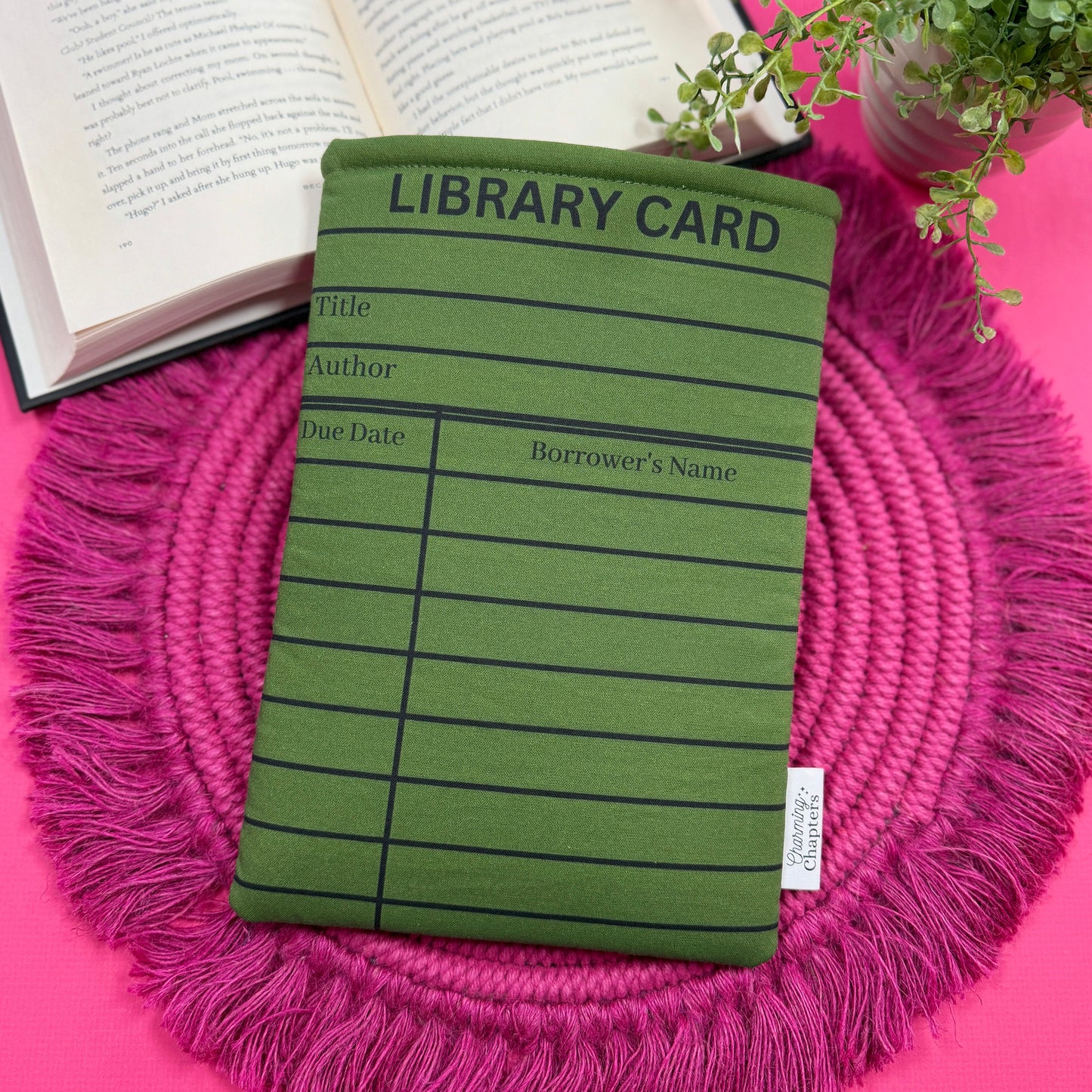 Padded book sleeve with a green and black library card design on a pink surface with an open book and plant in the background.
