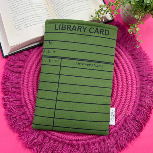 Padded book sleeve with a green and black library card design on a pink surface with an open book and plant in the background.