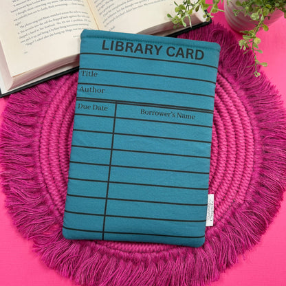 Padded book sleeve with a teal library card design on a pink surface with an open book and plant in the background.