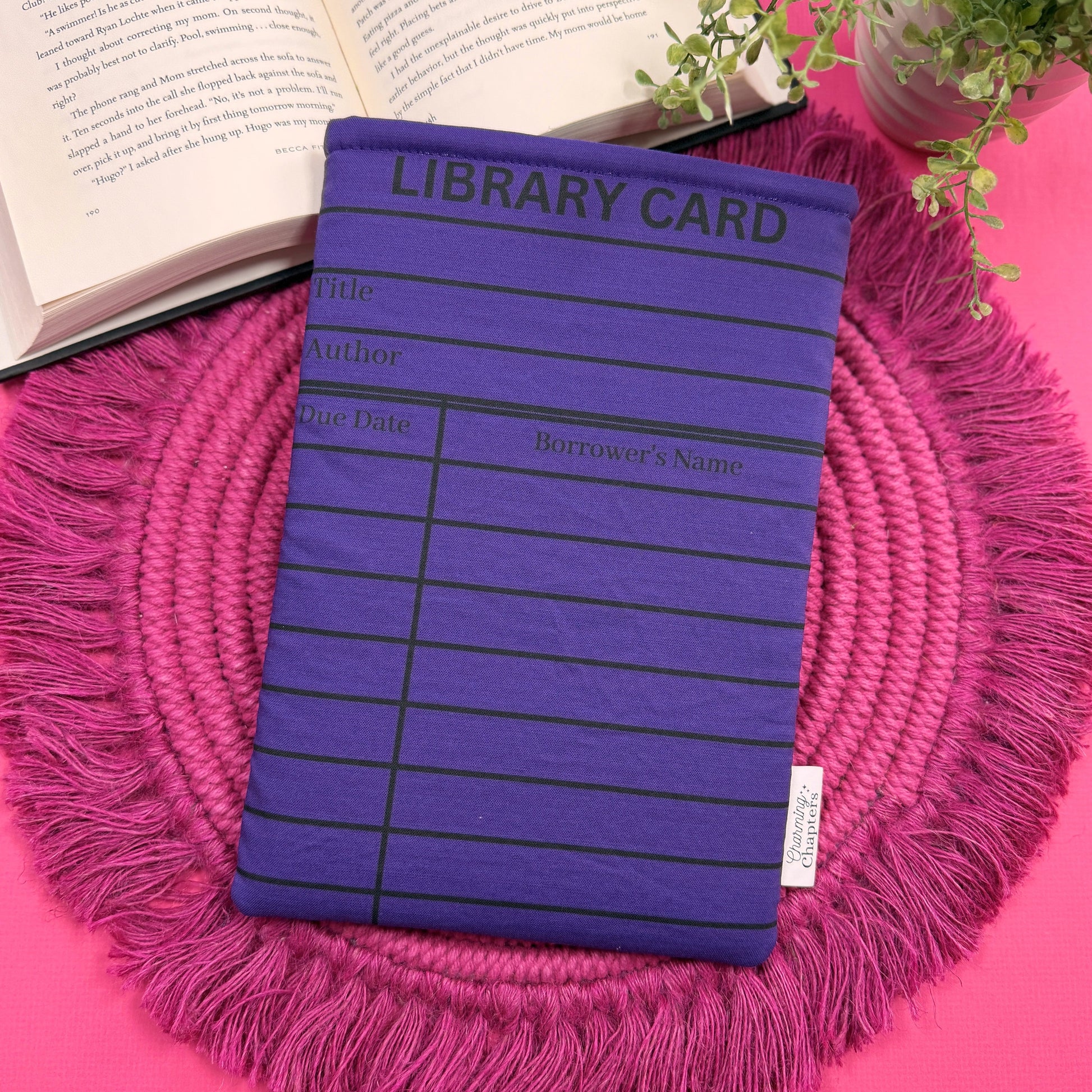 Padded book sleeve with a purple library card design on a pink surface with an open book and plant in the background.