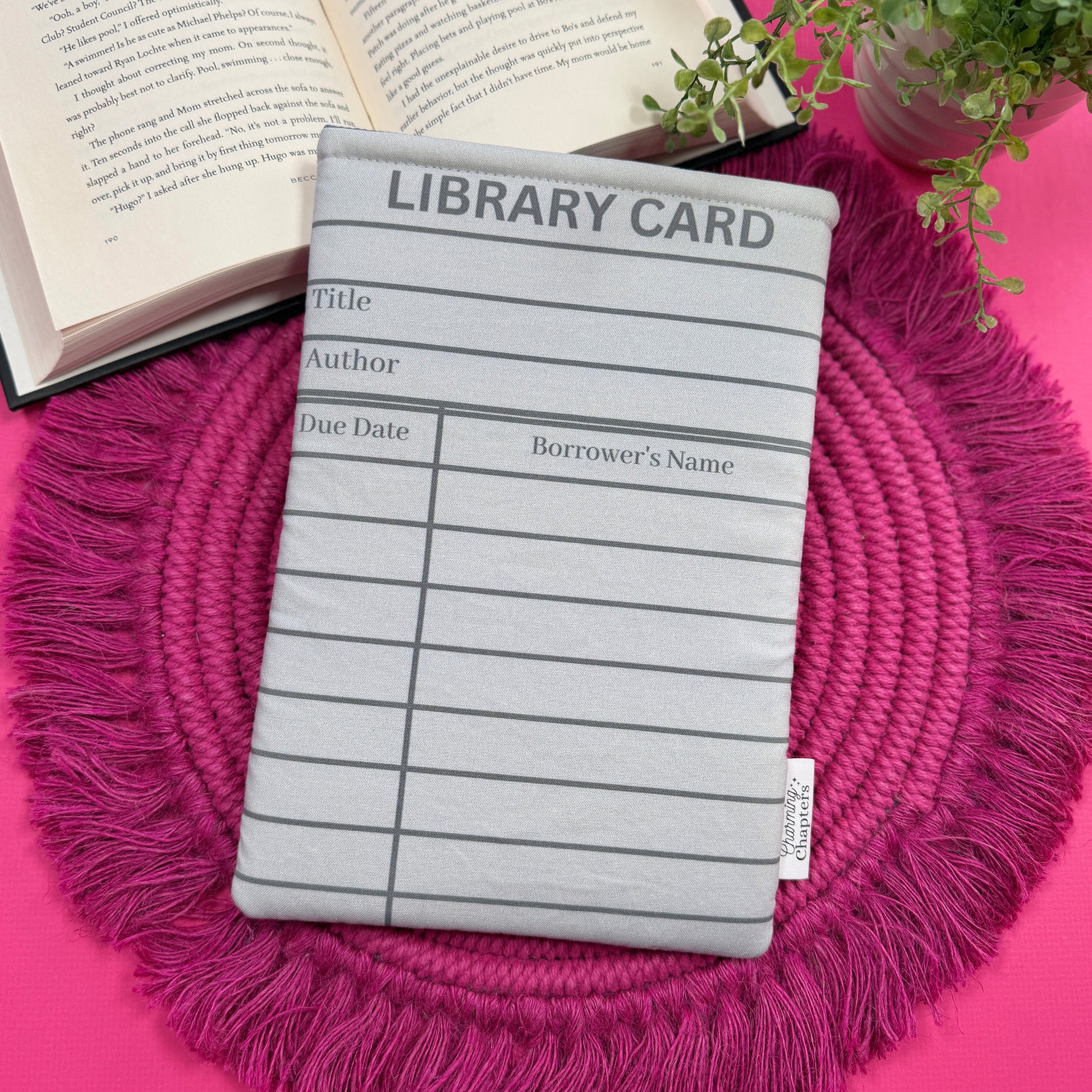 Padded book sleeve with a gray library card design on a pink surface with an open book and plant in the background.