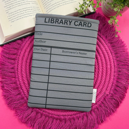 Padded book sleeve with a gray library card design on a pink surface with an open book and plant in the background.