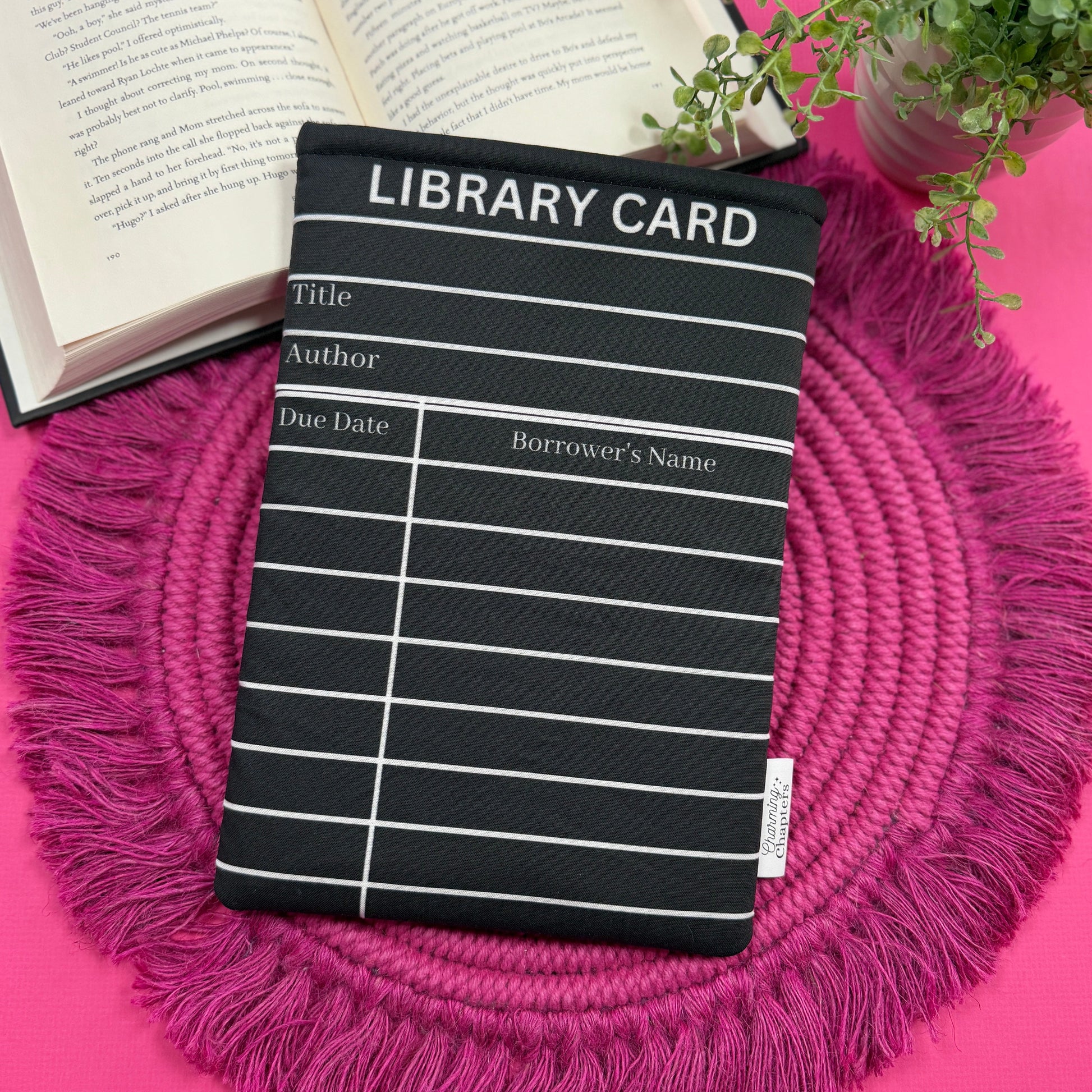 Padded book sleeve with a black and white library card design on a pink surface with an open book and plant in the background.