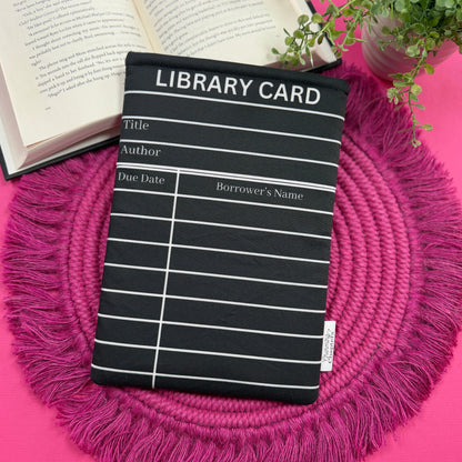 Padded book sleeve with a black and white library card design on a pink surface with an open book and plant in the background.