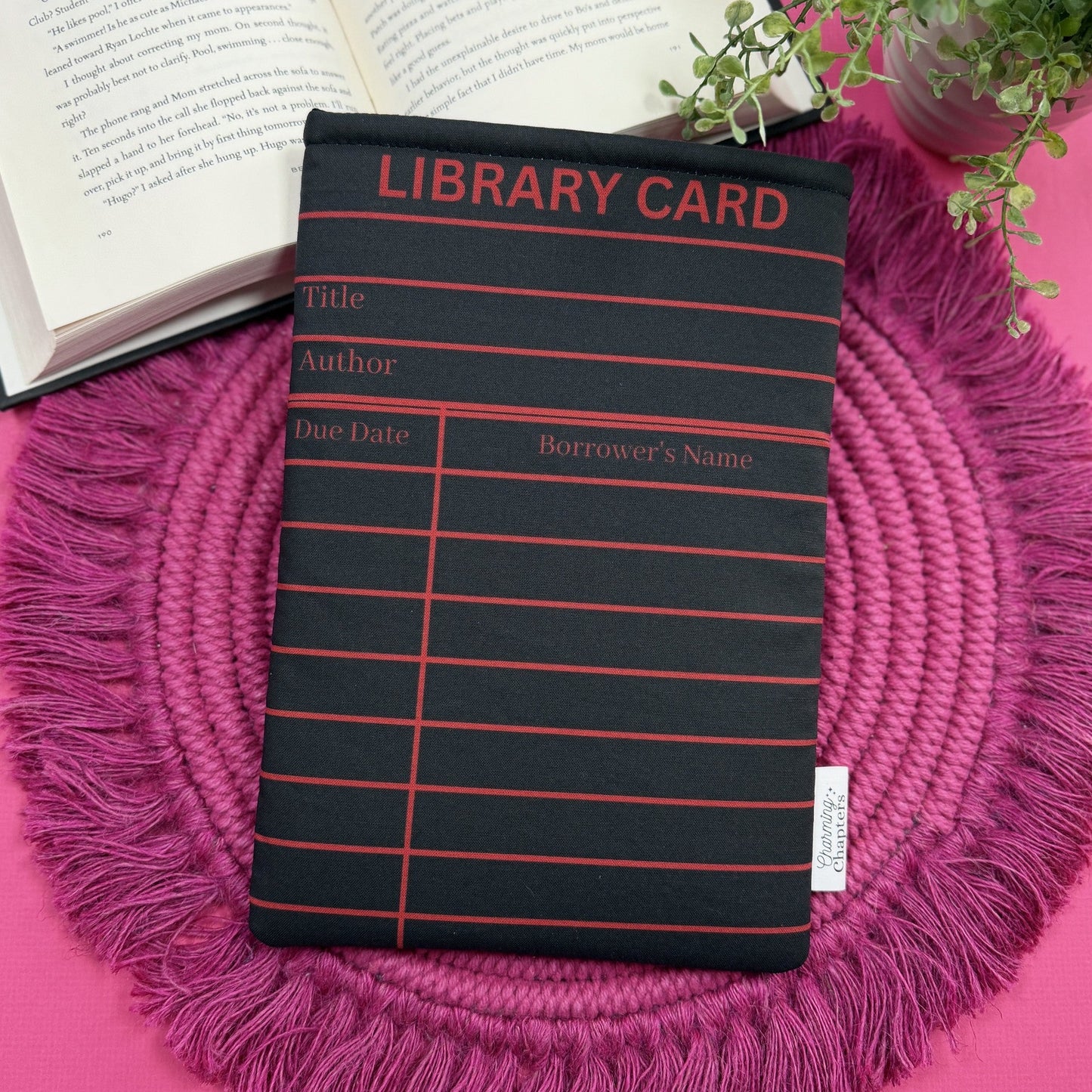 Padded book sleeve with a black and red library card design on a pink surface with an open book and plant in the background.