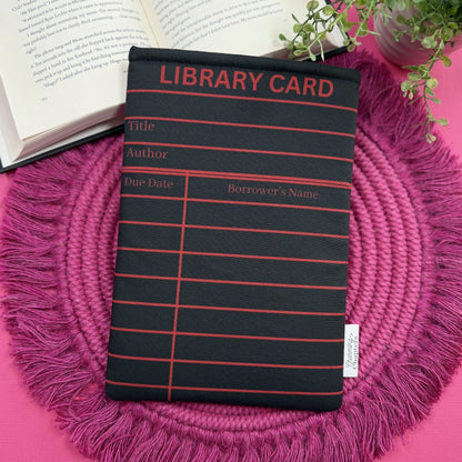 Padded book sleeve with a black and red library card design on a pink surface with an open book and plant in the background.