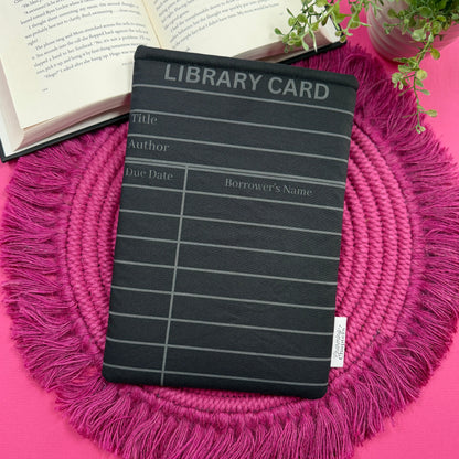 Padded book sleeve with a black and gray library card design on a pink surface with an open book and plant in the background.