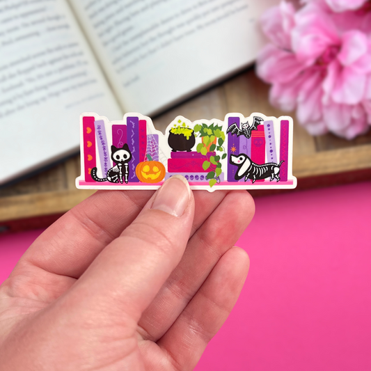 Hand holding a vinyl sticker with a Halloween bookshelf design, with an open book and pink flowers in the background.