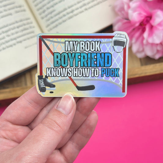 Hand holding a vinyl sticker with a hockey romance design, with an open book and pink flowers in the background. The sticker has a holographic finish and features a hockey stick, skates, net and helmet.
