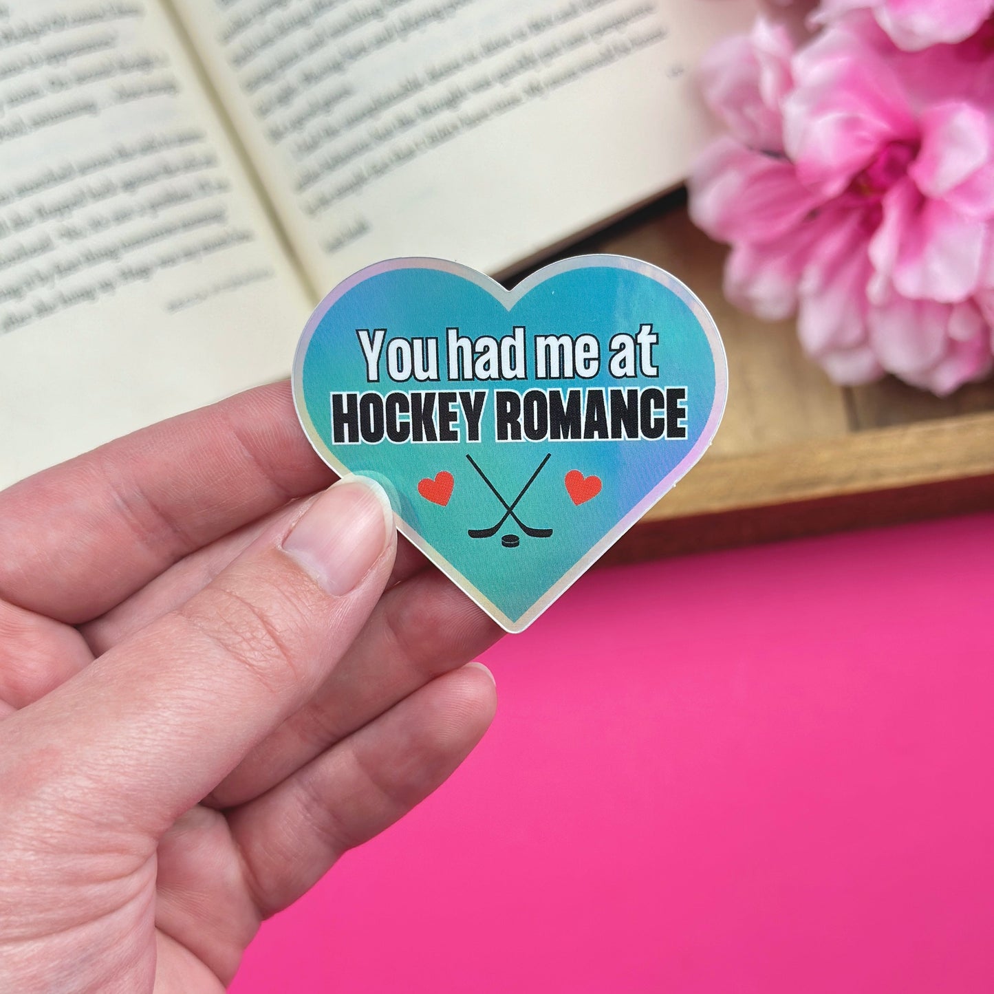 Hand holding a heart-shaped vinyl sticker with a hockey romance design, with an open book and pink flowers in the background. The sticker has a holographic finish and features hockey sticks and hearts.