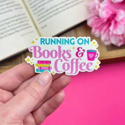 Hand holding a 'Running on Books & Coffee' sticker with an open book and pink flowers in the background.