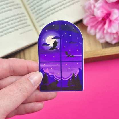 Vinyl sticker with a design that has a window showing dragons flying over mountains at night.  The sticker is being held by a hand on a pink surface.