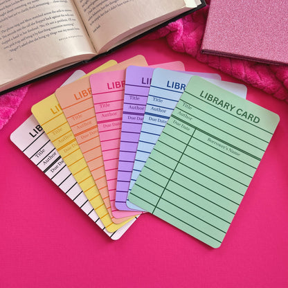 Colorful library card Kindle inserts laying on a pink surface with an open book.