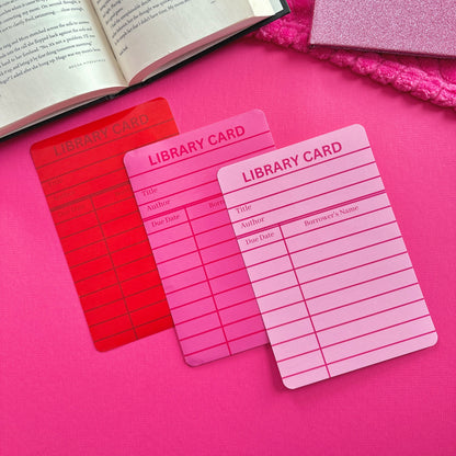 Three library card Kindle inserts laying on a pink surface with an open book.  One library card is red, one is dark pink, and one is light pink.