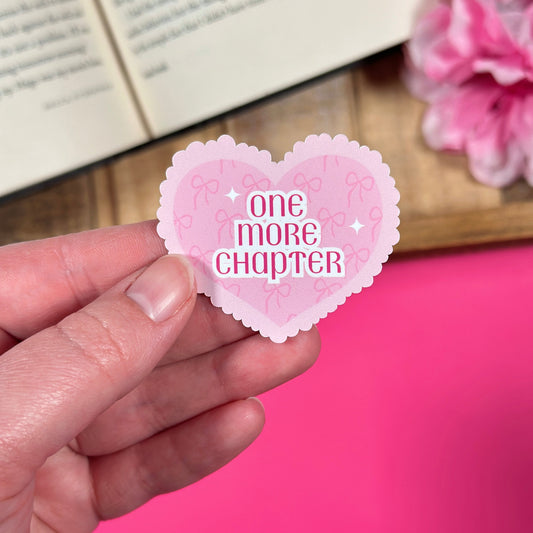 Heart-shaped sticker with 'one more chapter' text held by a hand against a pink background.