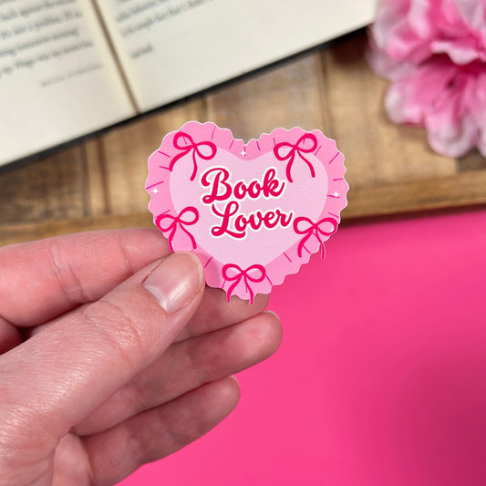 Heart-shaped 'Book Lover' sticker held by a hand with a book and pink flowers in the background.