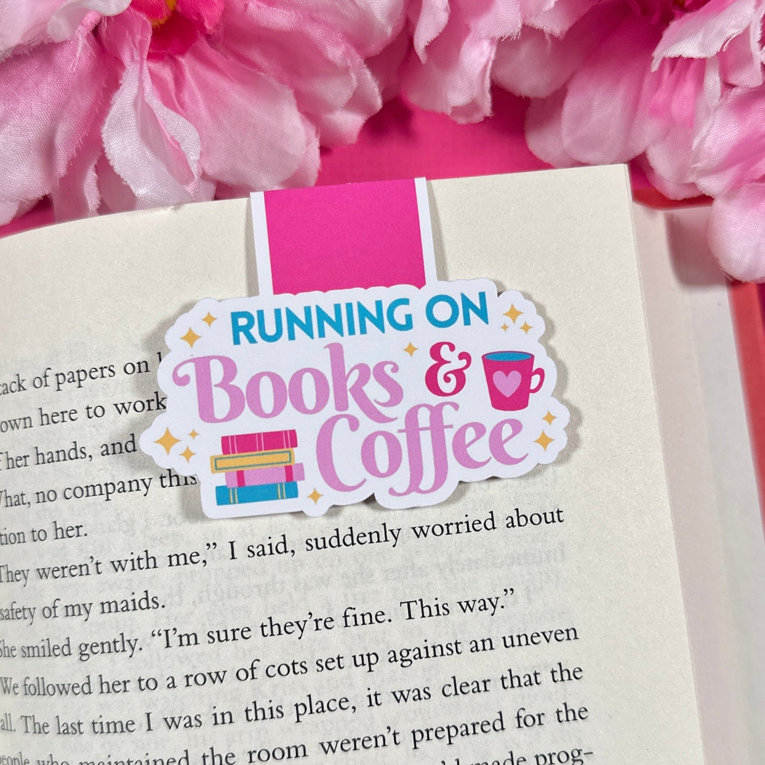 Open book with a 'Running on Books & Coffee' magnetic bookmark, surrounded by pink flowers.