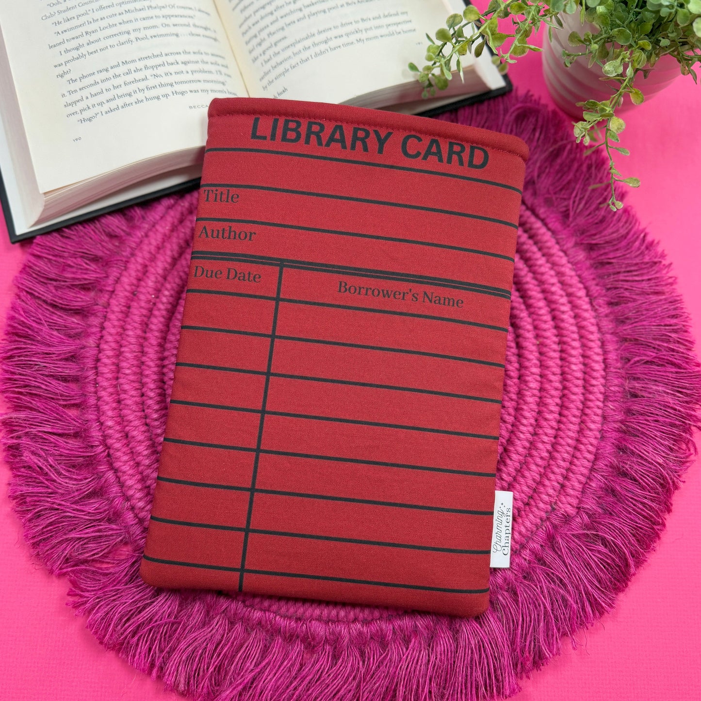 Padded book sleeve with a red and black library card design on a pink surface with an open book and plant in the background.