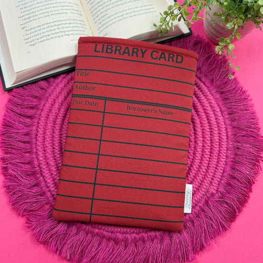 Padded book sleeve with a red and black library card design on a pink surface with an open book and plant in the background.