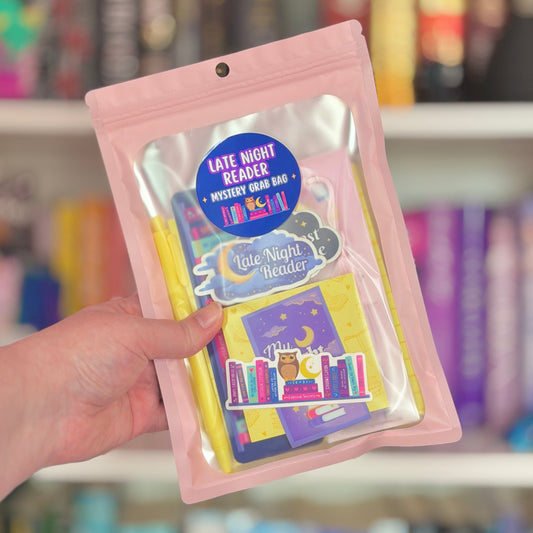 Hand holding a pink package with 'Late Night Reader Mystery Grab Bag' label against a blurred bookshelf background