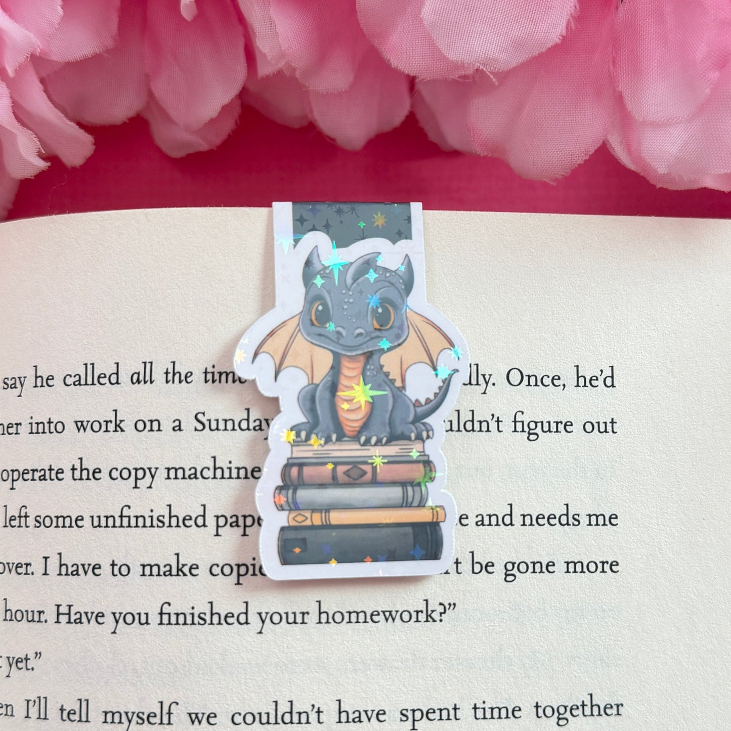 Magnetic bookmark with a black and golden yellow dragon design on an open book surrounded by pink flowers. The bookmark has a holographic star finish.