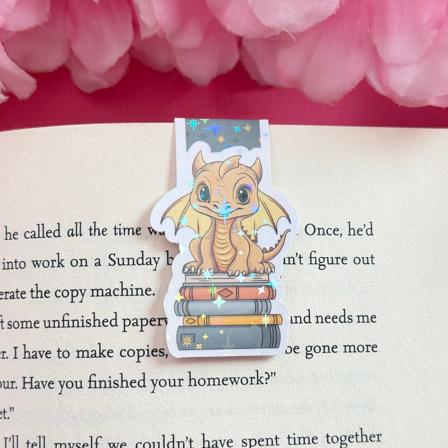 Magnetic bookmark with a golden yellow dragon design on an open book surrounded by pink flowers. The bookmark has a holographic star finish.