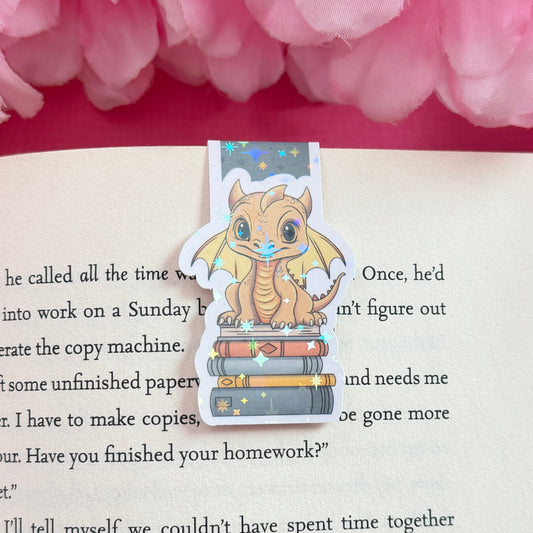 Magnetic bookmark with a golden yellow dragon design on an open book surrounded by pink flowers. The bookmark has a holographic star finish.